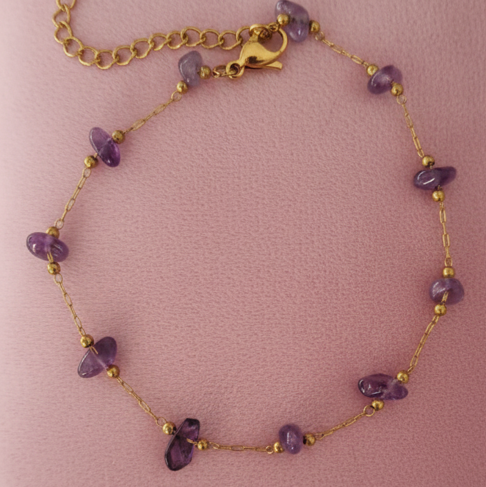 pulsera delicada de cadena dorada con piedras de amatista moradas naturales intercaladas. Pulsera de mujer minimalista en acero inoxidable con baño de oro 18k. Joyería espiritual y elegante sobre fondo rosa.